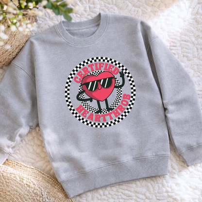 White Gildan youth sweatshirt flatlay with a Certified Heartthrob graphic across the front. The design features a smiling red heart character wearing sunglasses, framed by a black and white checkerboard circle and bold lettering. A fun choice for Valentine's Day school parties, family photos, holiday celebrations, or everyday wear during cooler weather.
