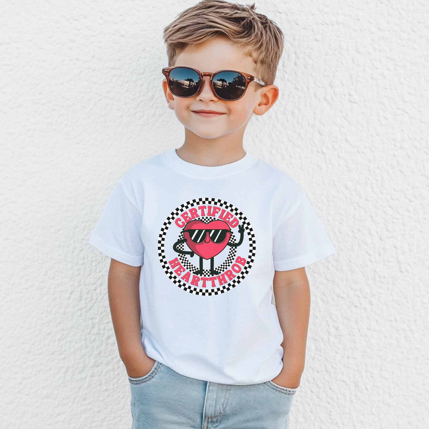 White youth Valentine's Day T shirt shown on a child, featuring a cheerful cartoon heart wearing sunglasses in the center of a black and white checkerboard circle. Certified Heartthrob text surrounds the graphic. The playful design is a fun choice for school celebrations, Valentine's parties, family photos, or everyday wear.