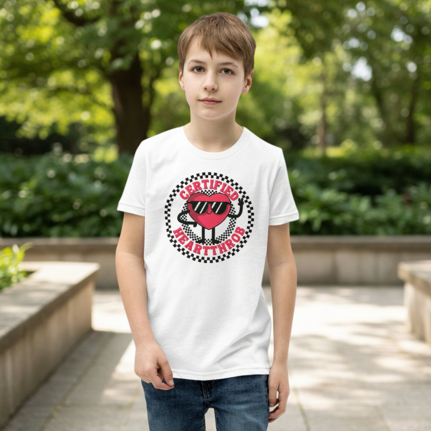 A child models a white youth graphic tee decorated with a playful Certified Heartthrob Valentine's Day design. A cartoon heart character wearing sunglasses sits inside a bold black and white checkerboard circle, with the phrase Certified Heartthrob surrounding the illustration. The fun design brings a cheerful personality to a child's holiday outfit.