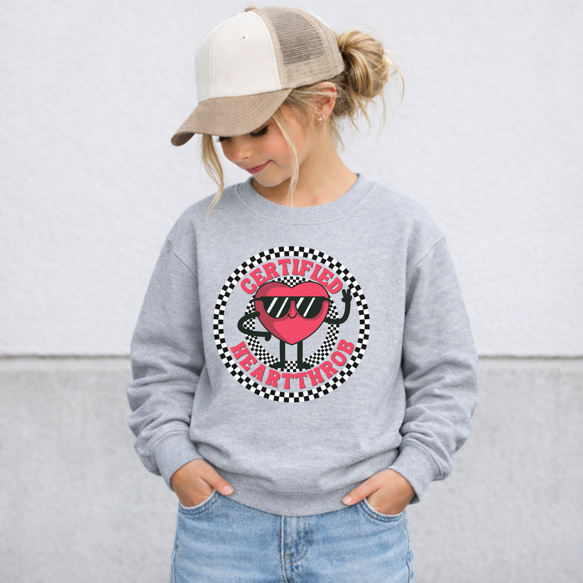 Gray youth Valentine's Day sweatshirt shown on a child, featuring a cheerful cartoon heart wearing sunglasses in the center of a black and white checkerboard circle. Certified Heartthrob text surrounds the graphic. The playful design is perfect for school celebrations, Valentine's parties, family photos, and chilly February days.