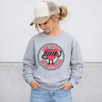 Gray youth Valentine's Day sweatshirt shown on a child, featuring a cheerful cartoon heart wearing sunglasses in the center of a black and white checkerboard circle. Certified Heartthrob text surrounds the graphic. The playful design is perfect for school celebrations, Valentine's parties, family photos, and chilly February days.