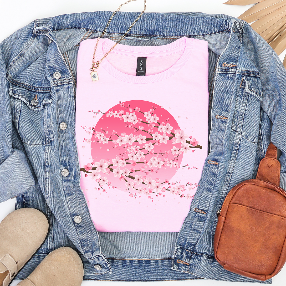 Folded light pink women's cherry blossom t shirt with pink sakura floral graphic, Japanese aesthetic design, styled with a denim jacket, gold necklace, brown crossbody bag and beige shoes