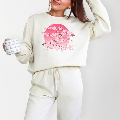Ivory Comfort Colors 1466 sweatshirt featuring a delicate cherry blossom design with pink sakura branches layered across a soft pink moon. The relaxed pullover has a cozy lived in feel and feminine artistic style, perfect for casual everyday outfits, coffee runs, school drop offs, and cool weather layering.