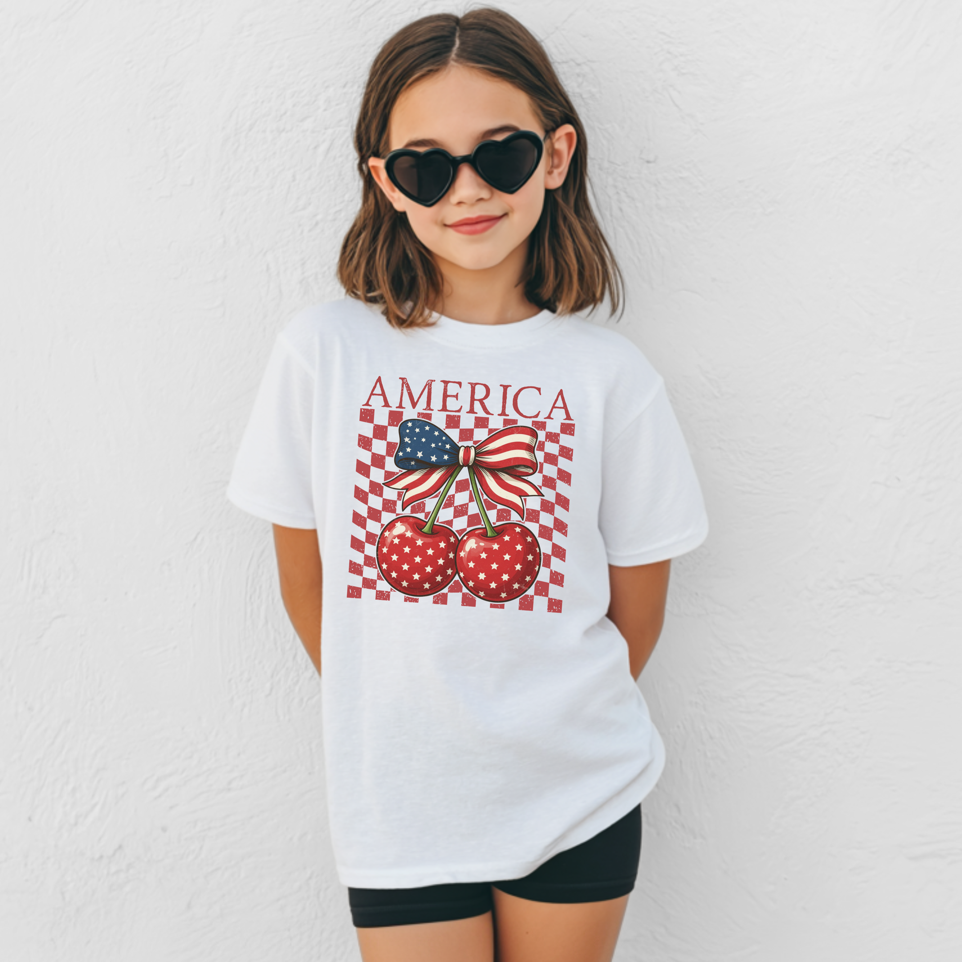 Girls patriotic Comfort Colors tee featuring America wording above a cherry bow design with red cherries, white star print, and a bold flag inspired ribbon. Distressed checkerboard print adds nostalgic Americana style to the front graphic. Youth garment dyed cotton shirt with classic fit and soft texture. Great gift for daughters, granddaughters, nieces, and little besties who need a cute Fourth of July shirt.