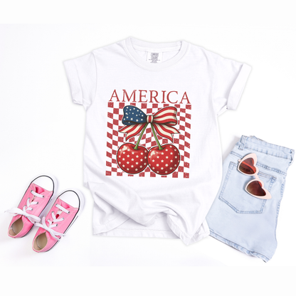 Youth patriotic graphic tee in white featuring America text with playful cherry bow artwork centered on the front. Two bright red cherries with white stars hang beneath a large American flag bow while faded checkerboard detail adds retro charm. Comfort Colors cotton shirt with soft washed feel and durable fit for everyday wear. Perfect girls Fourth of July outfit for cookouts, fireworks, lake trips, family gatherings, and mommy and me matching sets.