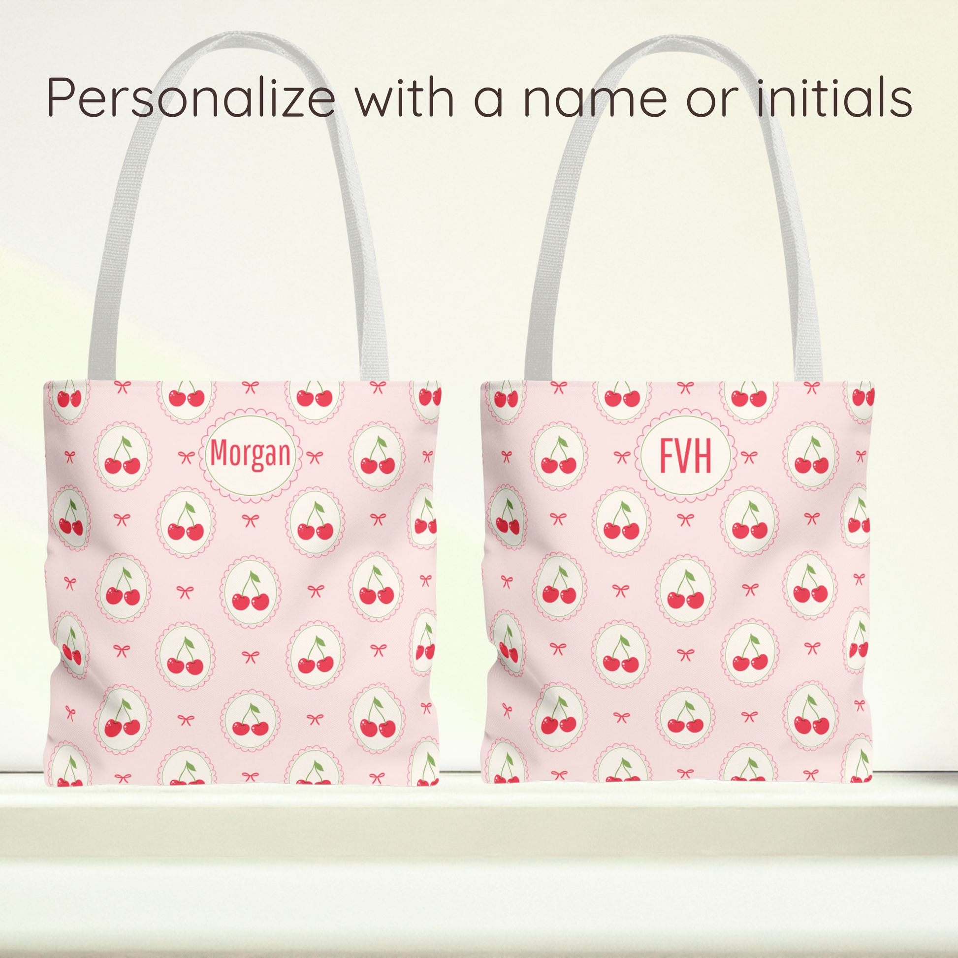 Two light pink tote bags with a repeating cherry print and white handles, shown side by side on a white surface against a pale background. Text at the top reads 'Personalize with a name or initials,' with the left bag personalized to 'Morgan' and the right bag to 'FVH'.