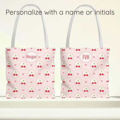 Two light pink tote bags with a repeating cherry print and white handles, shown side by side on a white surface against a pale background. Text at the top reads 'Personalize with a name or initials,' with the left bag personalized to 'Morgan' and the right bag to 'FVH'.