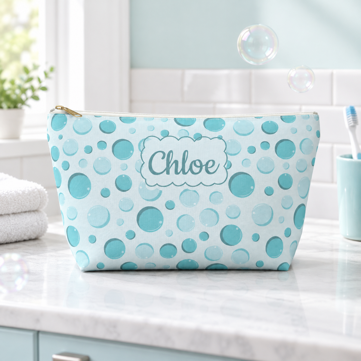 A custom cosmetic pouch with a light blue and teal bubble pattern sits on a white marble bathroom counter. The pouch is personalized with the name 'Chloe' in a cloud-shaped label.