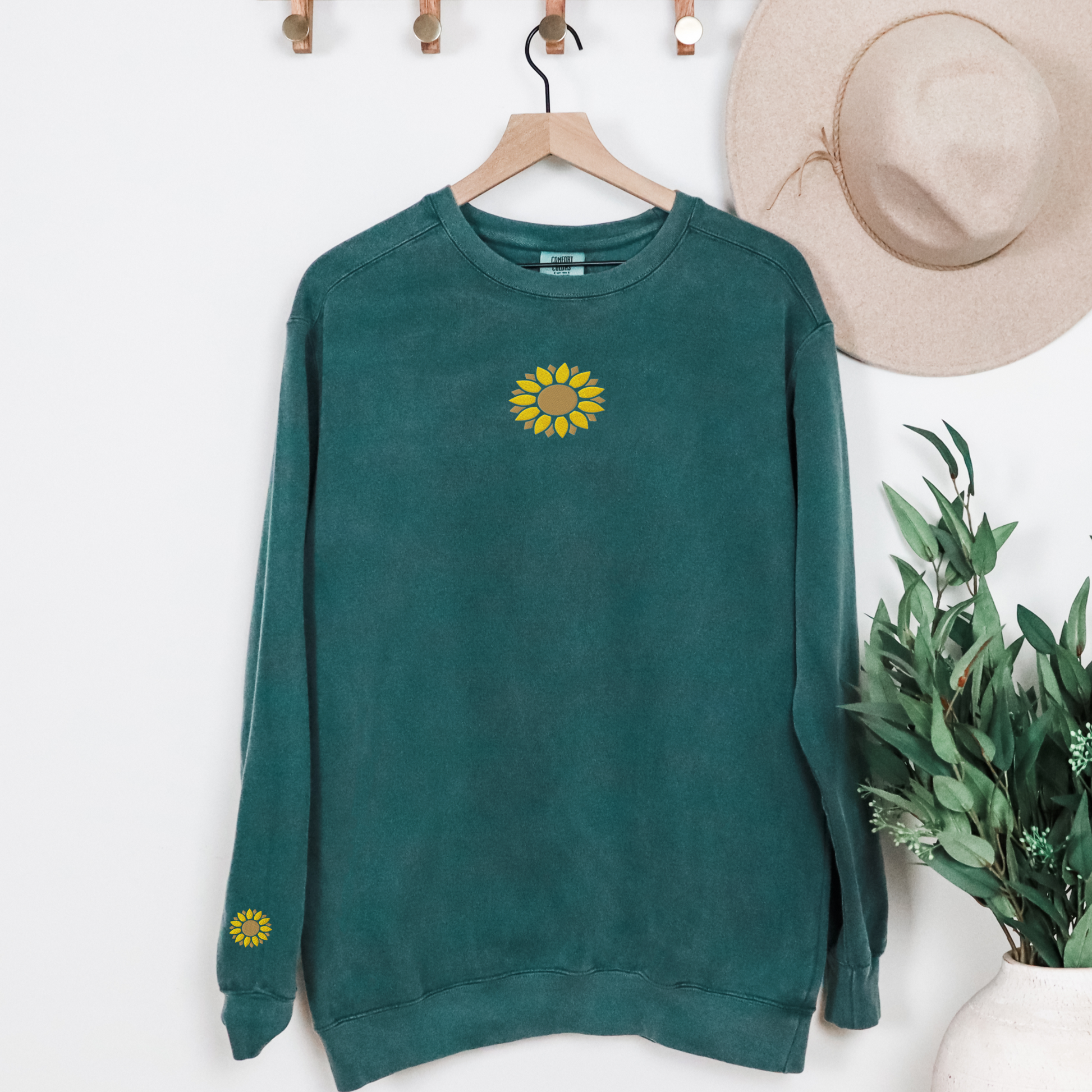 Blue Spruce Comfort Colors 1566 sweatshirt featuring embroidered sunflower details on the chest and right sleeve cuff. The garment dyed fabric creates a vintage inspired look while the floral embroidery adds a cheerful touch. This relaxed fit crewneck is ideal for nature lovers, gardening enthusiasts, and anyone searching for a comfortable floral sweatshirt to wear with jeans, leggings, or casual outfits.