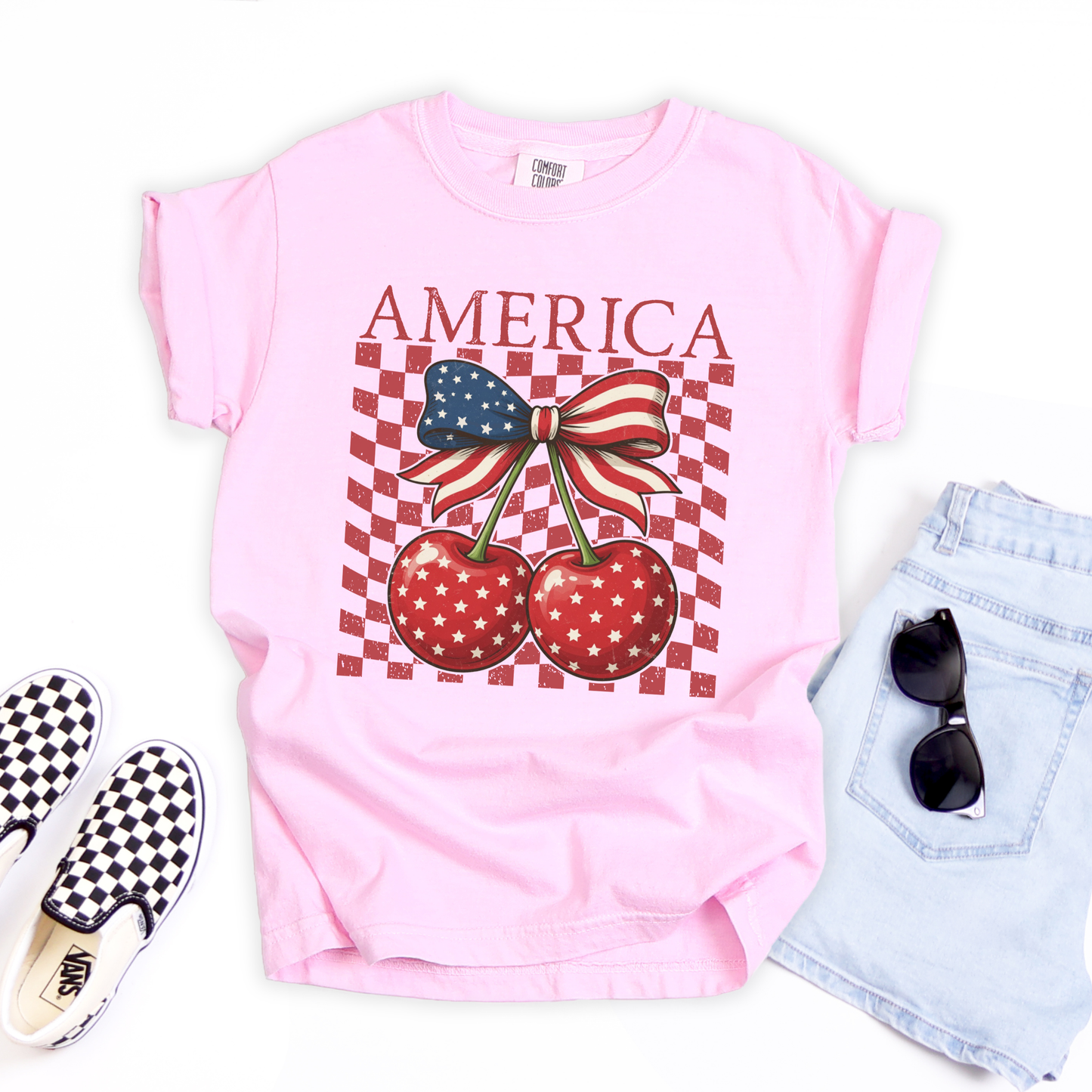 Girls Blossom Comfort Colors shirt with vintage America design and patriotic cherry bow graphic. Red cherries with white star pattern are tied together by a blue star and red stripe ribbon bow. Distressed checkerboard background creates trendy retro style. Garment dyed cotton youth tee with relaxed comfort for active kids. Great patriotic shirt for girls to wear to July fourth events, school parties, vacations, and matching family holiday outfits.