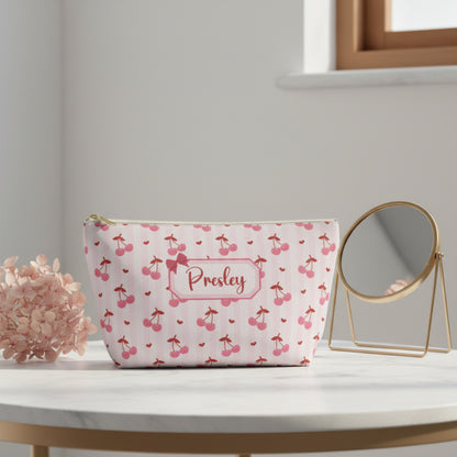Personalized cherry makeup bag with soft pink background, red cherry pattern, tiny heart details, and custom name label with bow design. Cute coquette cosmetic pouch featuring a white zipper with gold pull and roomy T bottom shape for makeup, skincare, toiletries, lip gloss, chargers, and travel essentials. Feminine soft girl aesthetic accessory bag perfect for teens, tweens, bridesmaids, birthday gifts, stocking stuffers, dance team gifts, and everyday purse organization with Free U.S. Shipping.