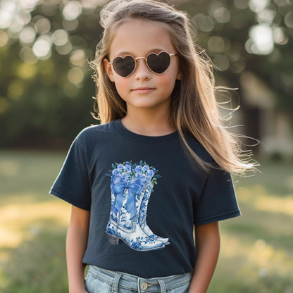A young girl with long blonde hair and heart-shaped sunglasses wears a navy blue t-shirt featuring a blue and white cowboy boot graphic filled with flowers, set against a blurred outdoor background.