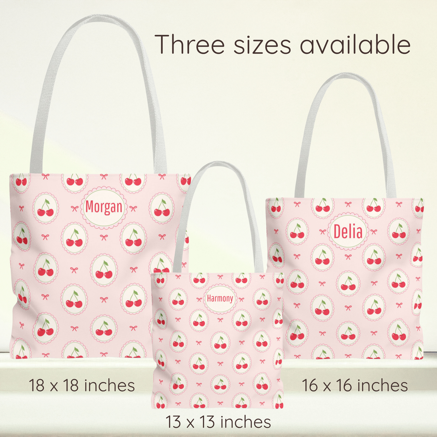 Three pink tote bags with white handles and a cherry print are shown in different sizes with text labels indicating 18 x 18 inches, 16 x 16 inches, and 13 x 13 inches. The bags have personalized names on the front: Morgan, Delia, and Harmony. Cute personalized cherry tote bag with a pale pink background and repeating cherry and bow pattern. The playful design combines cherry aesthetic, Kawaii, and Coquette style, making this reusable grocery bag a fun choice for back to school, farmers markets, beach days, 