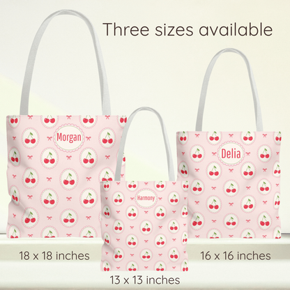 Three pink tote bags with white handles and a cherry print are shown in different sizes with text labels indicating 18 x 18 inches, 16 x 16 inches, and 13 x 13 inches. The bags have personalized names on the front: Morgan, Delia, and Harmony. Cute personalized cherry tote bag with a pale pink background and repeating cherry and bow pattern. The playful design combines cherry aesthetic, Kawaii, and Coquette style, making this reusable grocery bag a fun choice for back to school, farmers markets, beach days, 