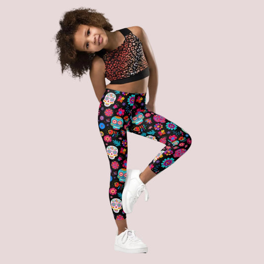 Young girl wearing colorful Dia de los Muertos leggings with a black background covered in vibrant sugar skulls, marigolds and colorful floral designs. The girls Day of the Dead leggings feature a bright all over print with pink, orange, yellow, blue, green and purple details, creating a festive look for Dia de los Muertos celebrations, cultural events, family photos and everyday wear.