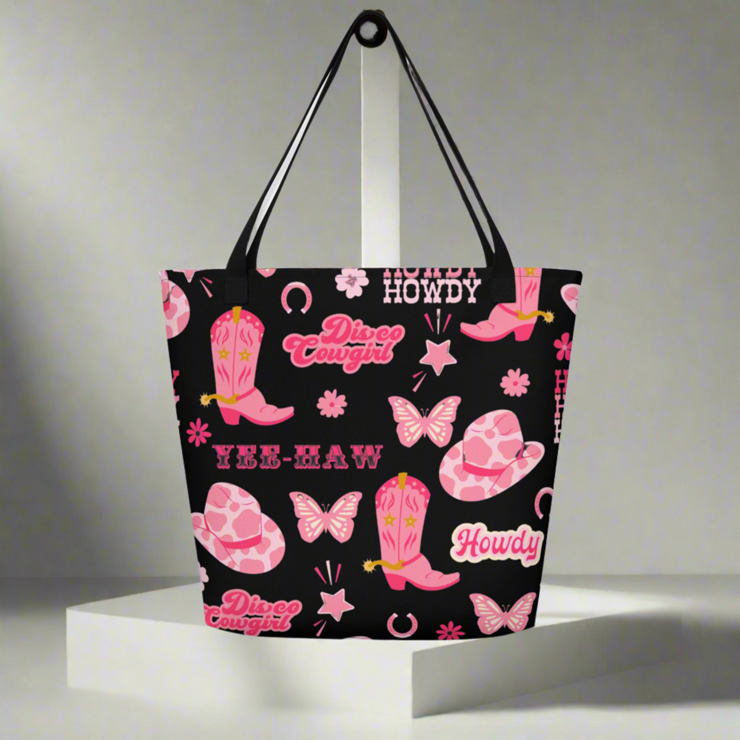 Image: Disco Cowgirl Tote Bag. This western glam canvas shoulder bag has a black background with a pink, white and pink pattern of butterflies, daisies, cowgirl hats and boots. The text on the bag reads Howdy, Yee-Haw and Disco Cowgirl. Great for weekend girls trips or bachelorette parties in Nashville, by jaecrece