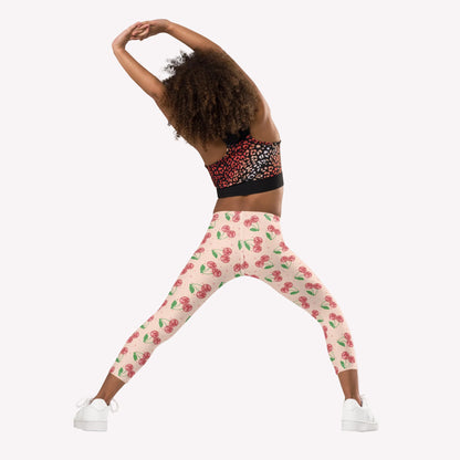 Girl wearing cherry print leggings in a soft pink tone with scattered red cherries, styled with a black and red athletic top and white sneakers. The leggings feature a high rise waistband and a full length fit that reaches the ankle. This outfit is ideal for active kids, whether heading to dance class, playing outside, or putting together a comfortable and stylish everyday look.