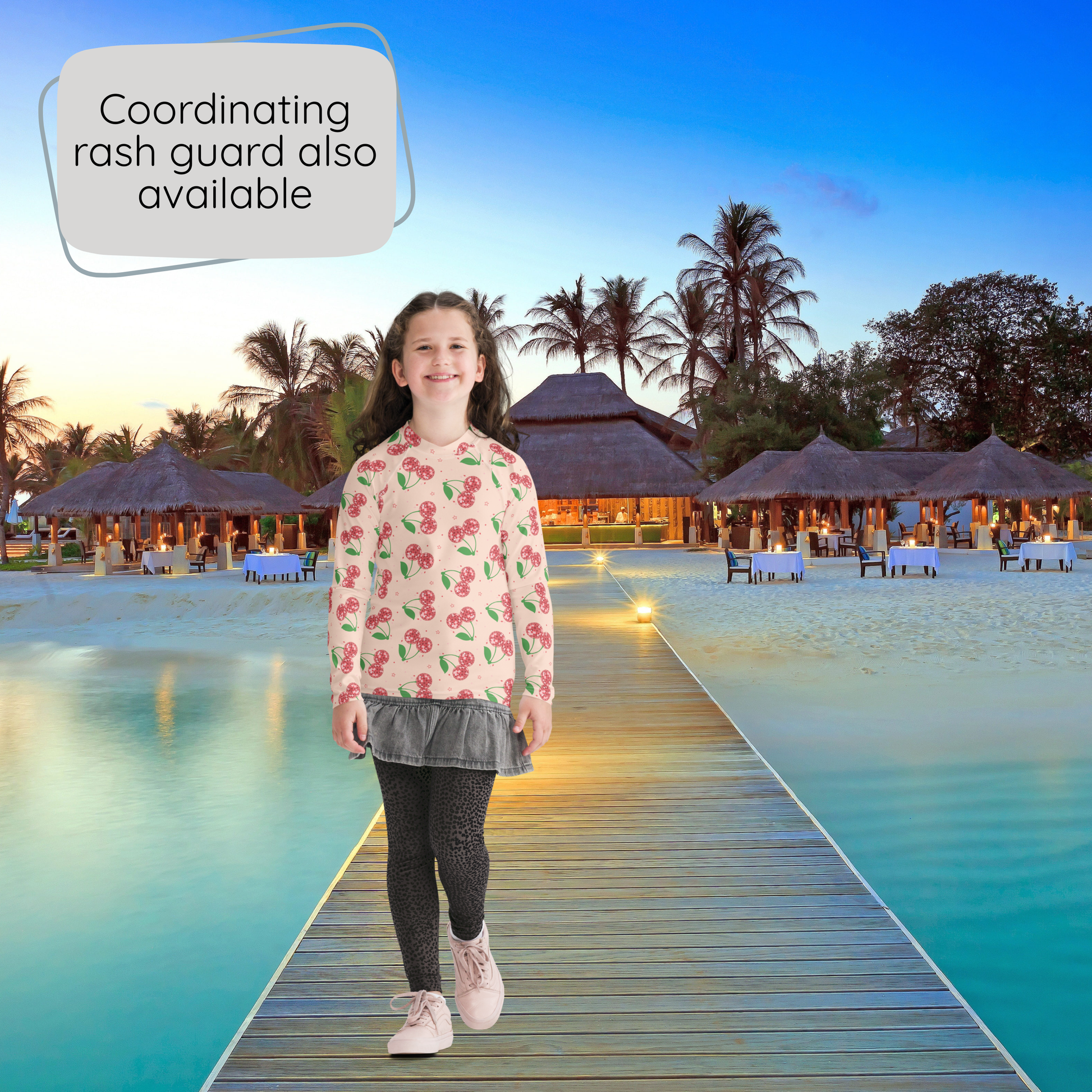 A young girl stands on a wooden boardwalk at a resort, wearing a pink long sleeve top with a green and red disco ball cherry pattern, a gray pleated skirt, black leggings, and pink sneakers. Palm trees, thatched umbrellas, and turquoise water are visible in the background, with a speech bubble in the top left reading 'Coordinating rash guard also available'.