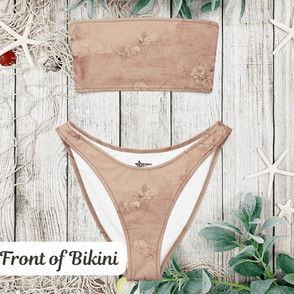 Flat lay of the Dusty Pink Cottagecore Bandeau Bikini top and bottom displayed on a rustic wooden beach-style surface. This soft pink, eco-friendly two-piece features a strapless bandeau top and high-leg V-cut bottoms. The romantic pastel aesthetic and sustainable recycled fabric make this swimsuit ideal for soft girl, coquette, and cottagecore summer looks. Available in inclusive sizing from 2XS–6XL.
