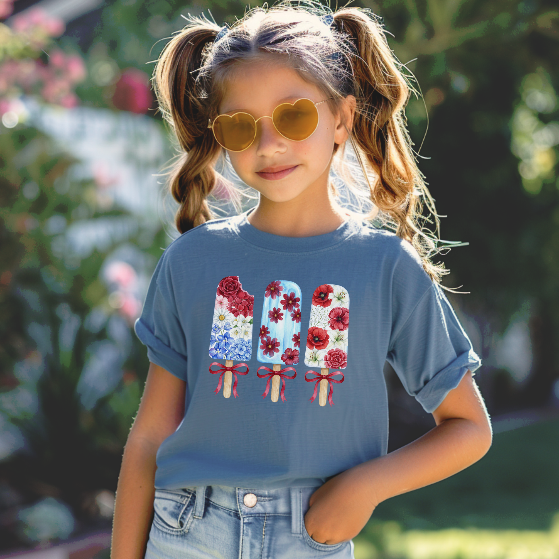A young girl with pigtails and heart shaped sunglasses wears a slate blue t-shirt featuring a graphic of three patriotic popsicles decorated with daisies and red roses, set against a blurred outdoor garden background.