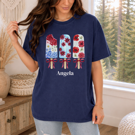 A woman wearing a navy blue t-shirt with a graphic of three popsicles decorated with red and blue floral patterns, and the name 'Angela' printed below. She is standing in a bright room with a window showing a forest view.