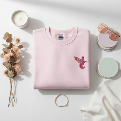 Folded light pink Gildan crewneck sweatshirt featuring a colorful embroidered hummingbird design on the chest. The neatly folded sweatshirt highlights the soft pink fabric and detailed bird embroidery while giving shoppers a clear look at the garment color and overall style. This nature inspired sweatshirt is a comfortable everyday choice for hummingbird lovers, bird lovers, gardeners, and anyone who enjoys subtle embroidered clothing.