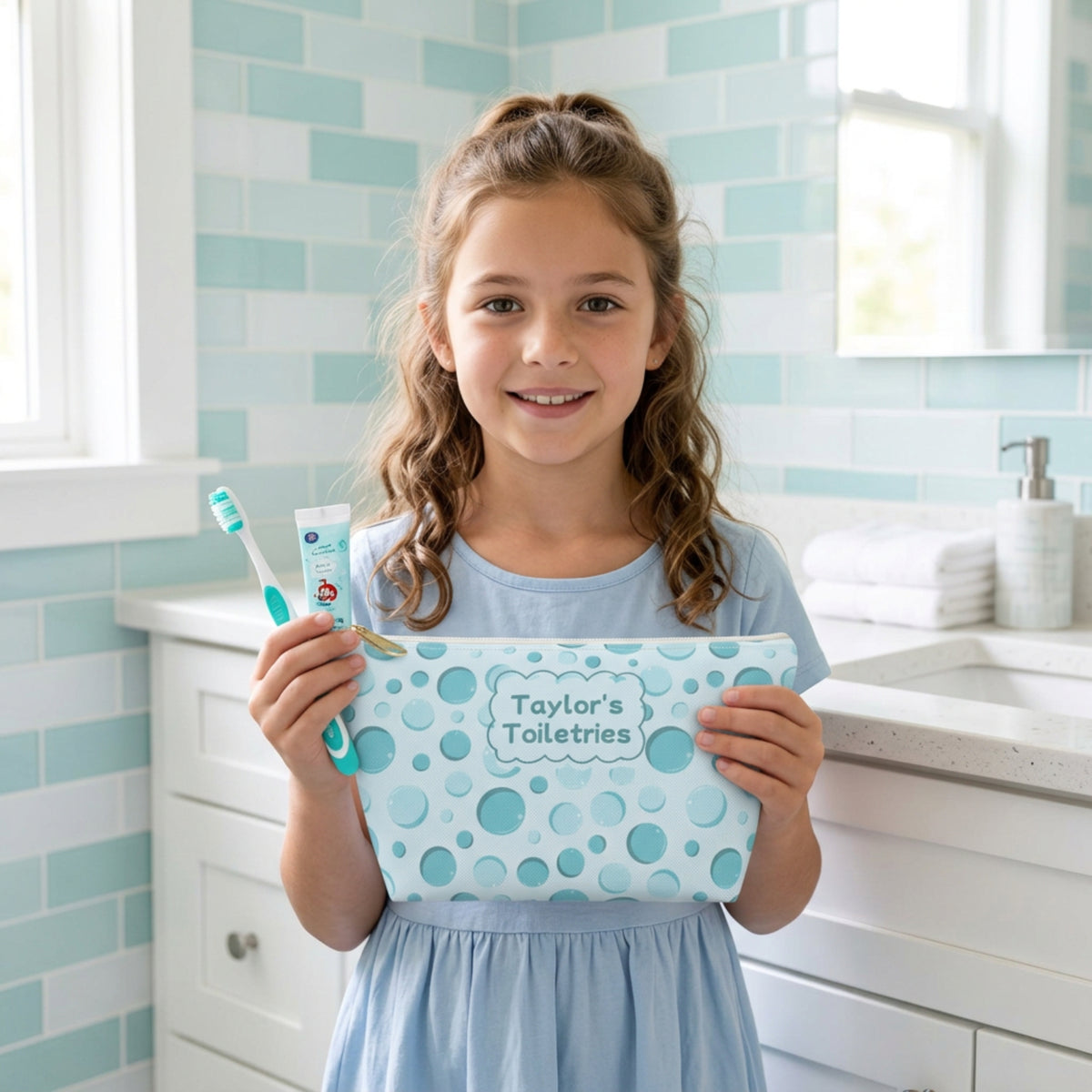 School aged girl holding a personalized aqua bubble print toiletry pouch alongside a children's toothbrush and toothpaste in a bright bathroom. The compact zippered bag features a colorful bubble pattern and custom name label, making it a cheerful travel accessory for kids to use for sleepovers, summer camp, and family trips.