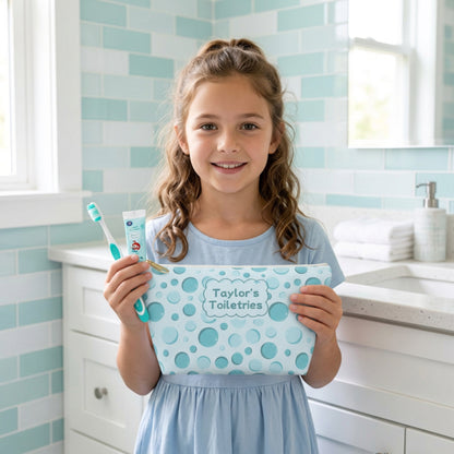 School aged girl holding a personalized aqua bubble print toiletry pouch alongside a children's toothbrush and toothpaste in a bright bathroom. The compact zippered bag features a colorful bubble pattern and custom name label, making it a cheerful travel accessory for kids to use for sleepovers, summer camp, and family trips.