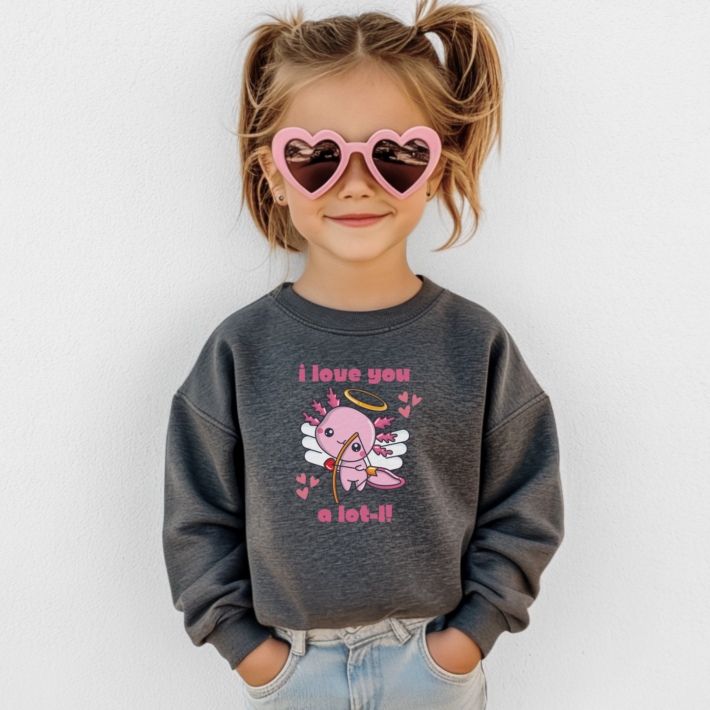 A girl wearing a heather grey youth Valentine sweatshirt with a sweet axolotl cupid design, heart accents, and the phrase I love you a lot l. The neutral gray tone pairs easily with everyday outfits while the classic crewneck fit keeps kids comfortable and warm. A cute and wearable Valentine sweatshirt for school, casual wear, and matching family Valentine outfits.