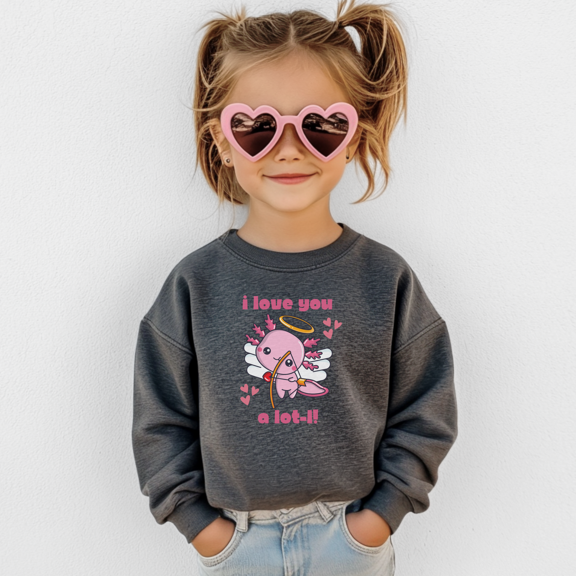 A girl wearing a heather grey youth Valentine sweatshirt with a sweet axolotl cupid design, heart accents, and the phrase I love you a lot l. The neutral gray tone pairs easily with everyday outfits while the classic crewneck fit keeps kids comfortable and warm. A cute and wearable Valentine sweatshirt for school, casual wear, and matching family Valentine outfits.