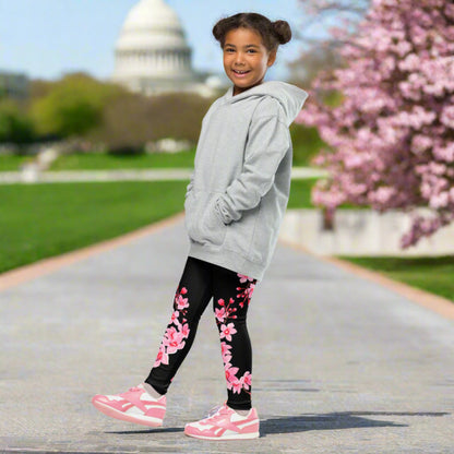 Young girl wearing black full length leggings with a delicate pink Japanese cherry blossom pattern while walking along a sidewalk in Washington DC. The high rise girls leggings combine a soft stretchy feel with a feminine sakura floral design, making them a comfortable choice for spring outings, sightseeing, family trips, cherry blossom season, school days, and everyday adventures.