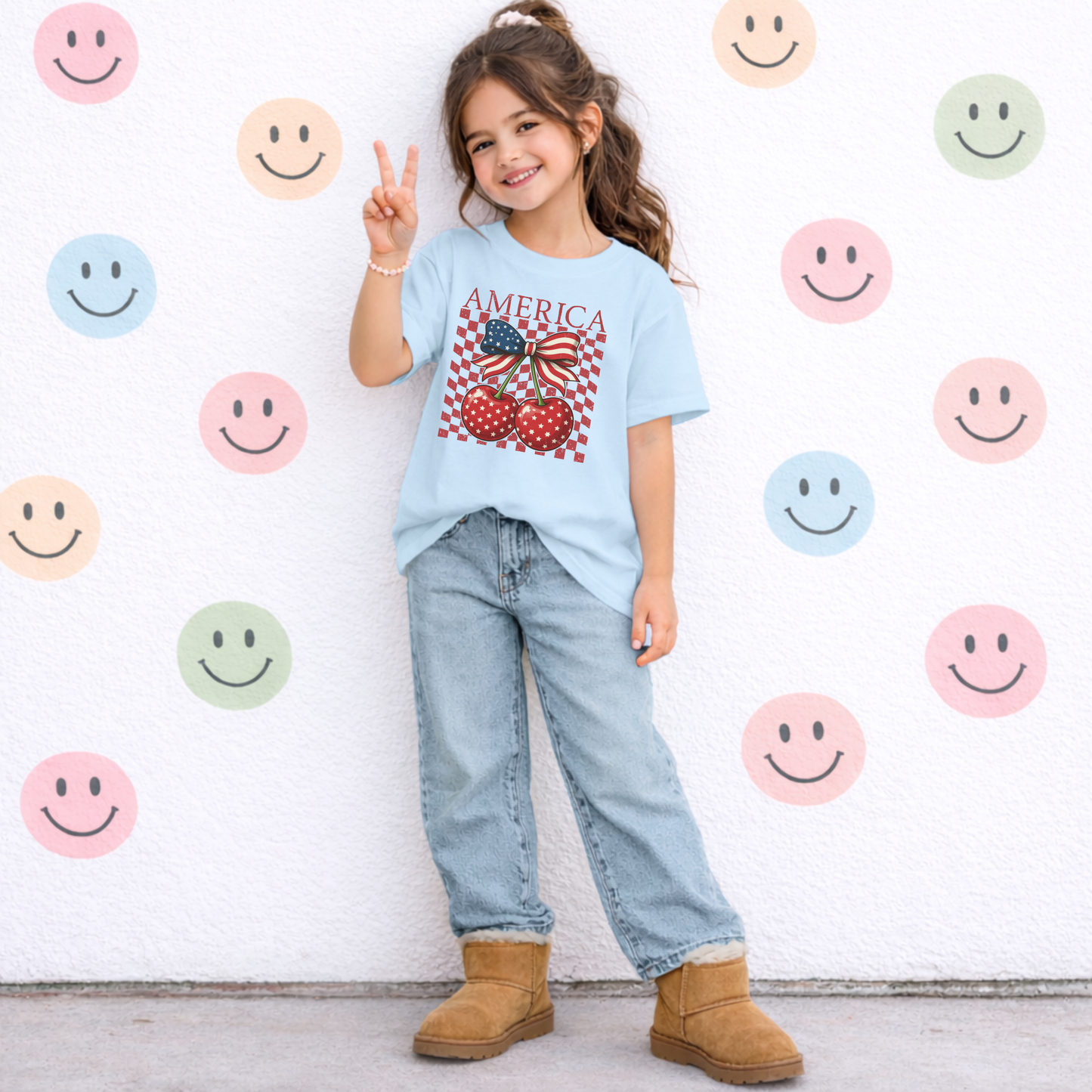 Girls Comfort Colors patriotic shirt with large America lettering and playful cherry coquette graphic. Red glossy cherries with white stars sit under a bold American flag bow while checkerboard accents create vintage race inspired flair. Garment dyed youth cotton tee offers comfortable all day wear and durable quality. Cute outfit choice for little girls during fireworks shows, summer parties, vacations, and coordinated family celebrations.