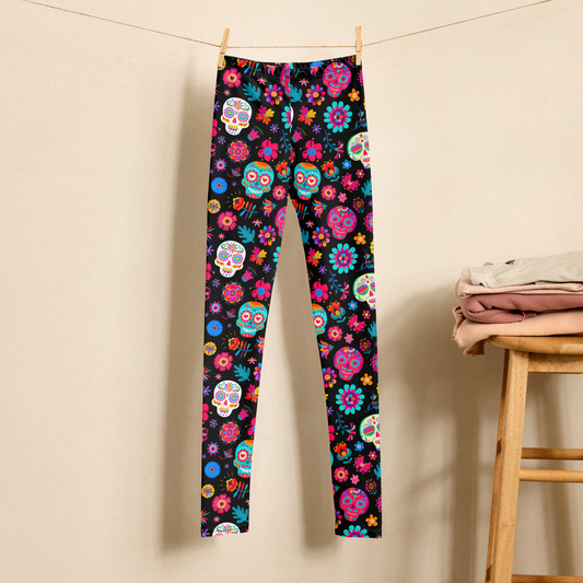 Girls Día de los Muertos leggings featuring colorful sugar skulls, flowers, butterflies, and festive floral details on a black background. The high waist, full length leggings are shown hanging in a neutral bedroom setting. Available in toddler, tween, and teen girls sizes 2T through 20, with matching women’s sizes available for Mommy and Me outfits.