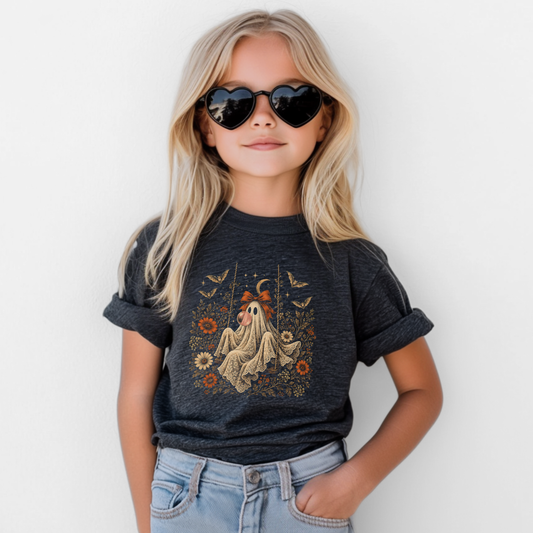 Girl wearing a dark heather grey Halloween T shirt featuring a whimsical ghost blowing bubble gum, surrounded by orange flowers, botanical foliage, bats, stars, lace and a bow. A vintage inspired girls graphic tee for spooky season, Halloween parties, fall activities and everyday autumn outfits.