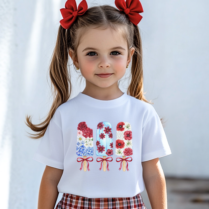 A young girl with brown pigtails and red bows wears a white short-sleeve T-shirt featuring a colorful graphic of three floral patriotic popsicles, paired with a red and white plaid skirt against a bright, softly blurred background.