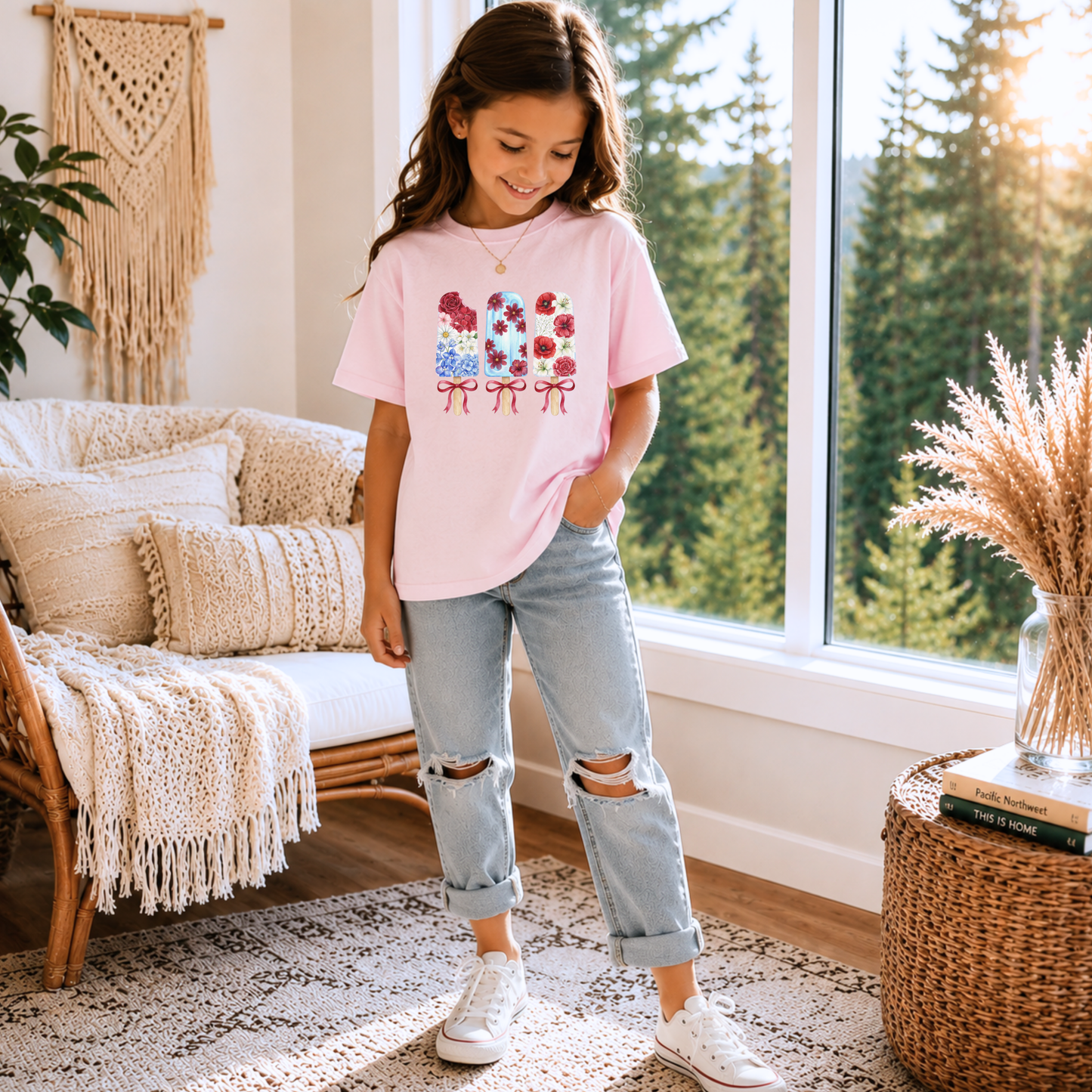 A young girl stands in a bright room wearing a light pink graphic T-shirt with a floral bow design, light-wash ripped jeans, and white sneakers.