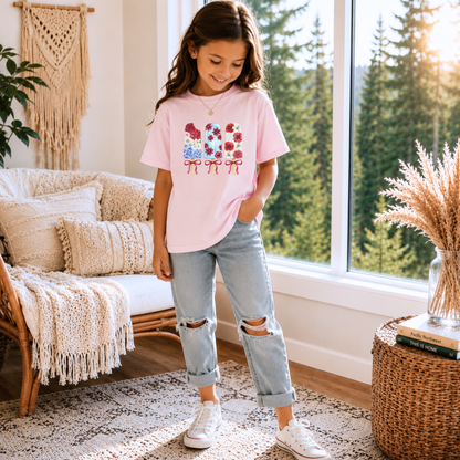 A young girl stands in a bright room wearing a light pink graphic T-shirt with a floral bow design, light-wash ripped jeans, and white sneakers.