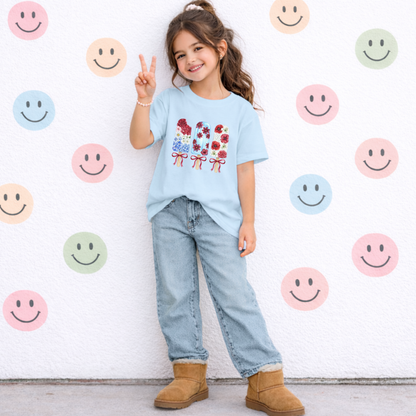A young girl stands against a white wall decorated with colorful smiley face stickers, wearing a light blue graphic t-shirt, light-wash jeans, and tan suede boots.