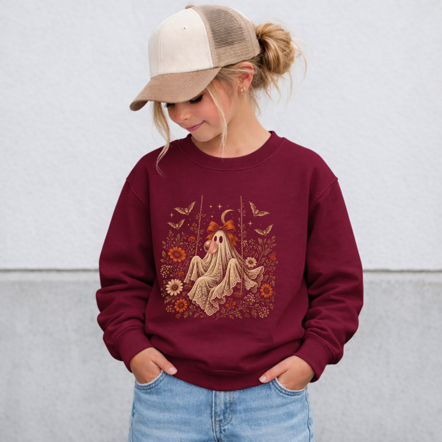 Girl wearing a maroon Halloween sweatshirt featuring a whimsical vintage ghost blowing bubble gum, surrounded by orange flowers, botanical details, bats, stars, lace and a bow. This cozy girls Halloween outfit combines vintage artwork with a playful spooky season aesthetic for fall.