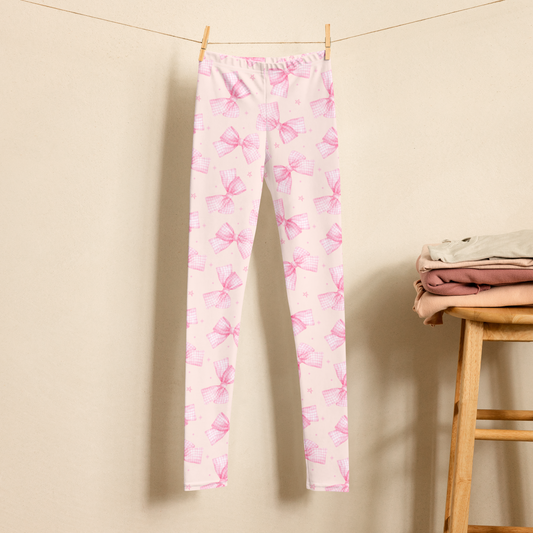 Girls pink bow leggings featuring a soft pink and white gingham bow pattern with delicate stars on a pale pink background. The high waist, full length leggings are shown hanging in a neutral setting and are available in sizes 2T through 20 for toddlers, tweens, and teens. Matching women’s sizes are also available for coordinated Mommy and Me outfits.