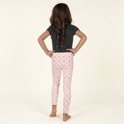 Back view of a young girl wearing pink heart leggings featuring a soft light pink background, delicate diamond lattice pattern, and scattered pink hearts. These full length girls leggings extend to the ankles and feature a comfortable high rise elastic waistband. Made from stretchy polyester and spandex fabric, these toddler and kids leggings are available in sizes 2T through 7 and are perfect for everyday outfits, school, playdates, and Valentine's Day celebrations.