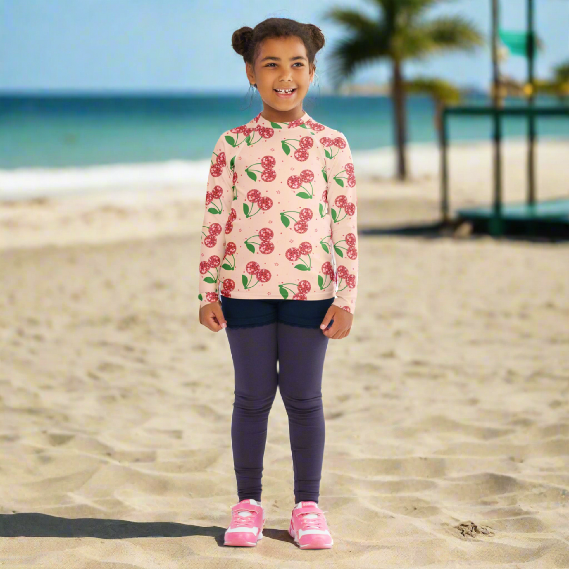 Girls long sleeve rash guard featuring a pink disco cherry pattern with red cherries on a soft pastel background. Available in toddler sizes 2T to 7, this fitted swim top provides UPF 50 sun protection and a comfortable stretch fit. Ideal for active kids at the beach, pool, or on vacation. A cute and playful swimwear option that combines style and function, with matching mommy and me swimwear available for family outfit sets.