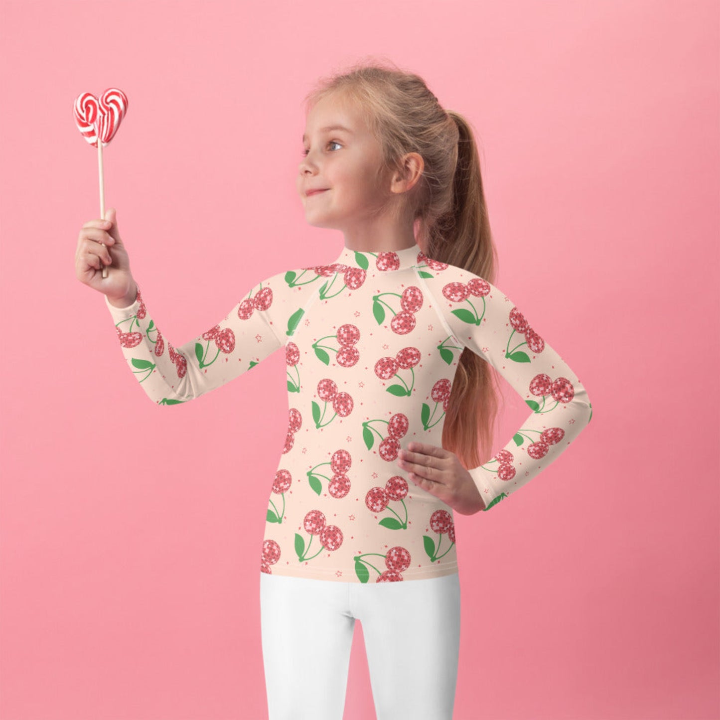 Girls disco cherry rash guard in toddler sizes 2T to 7 featuring a soft pink background with vibrant red cherry print. Modeled by a young girl in a bright swim setting, highlighting the long sleeve design and fitted silhouette. This girls swim top offers UPF 50 sun protection and comfortable stretch fabric for all day wear. Perfect for beach trips, pool days, vacations, and splash pad fun, plus available in matching mommy and me swimwear for coordinated family outfits.