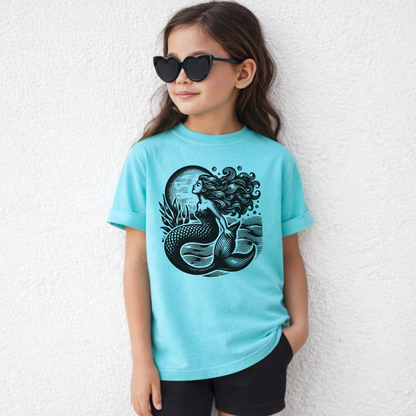 Girls mermaid graphic t shirt featuring a linocut inspired ocean illustration with a mermaid rising from textured sea waves beneath a glowing full moon. Printed on a soft Comfort Colors youth tee with a relaxed fit and vintage garment dyed finish. Perfect coastal aesthetic shirt for beach vacations summer outfits seaside adventures and matching mommy and me looks. Ideal for ocean loving girls who love whimsical nautical fashion mermaidcore style and comfy everyday graphic tees.