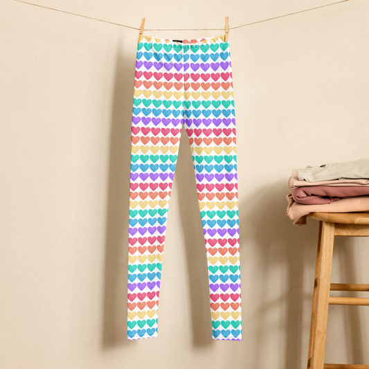 Girls rainbow heart leggings hanging against a light background, featuring rows of colorful hearts in a cheerful rainbow pattern. These high waist, full length leggings are available in sizes 2T through 20 for toddlers, tweens, and teens, making them fun for Pride celebrations or everyday wear. Matching women’s sizes are also available for a coordinated Mommy and Me look.
