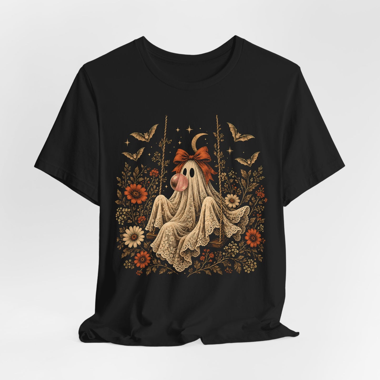 Gothic cottagecore inspired Halloween shirt featuring a vintage ghost sitting on a swing while blowing bubble gum. Floral and botanical details surround the ghost along with bats, lace, stars and a crescent moon for a romantic whimsical spooky season look.