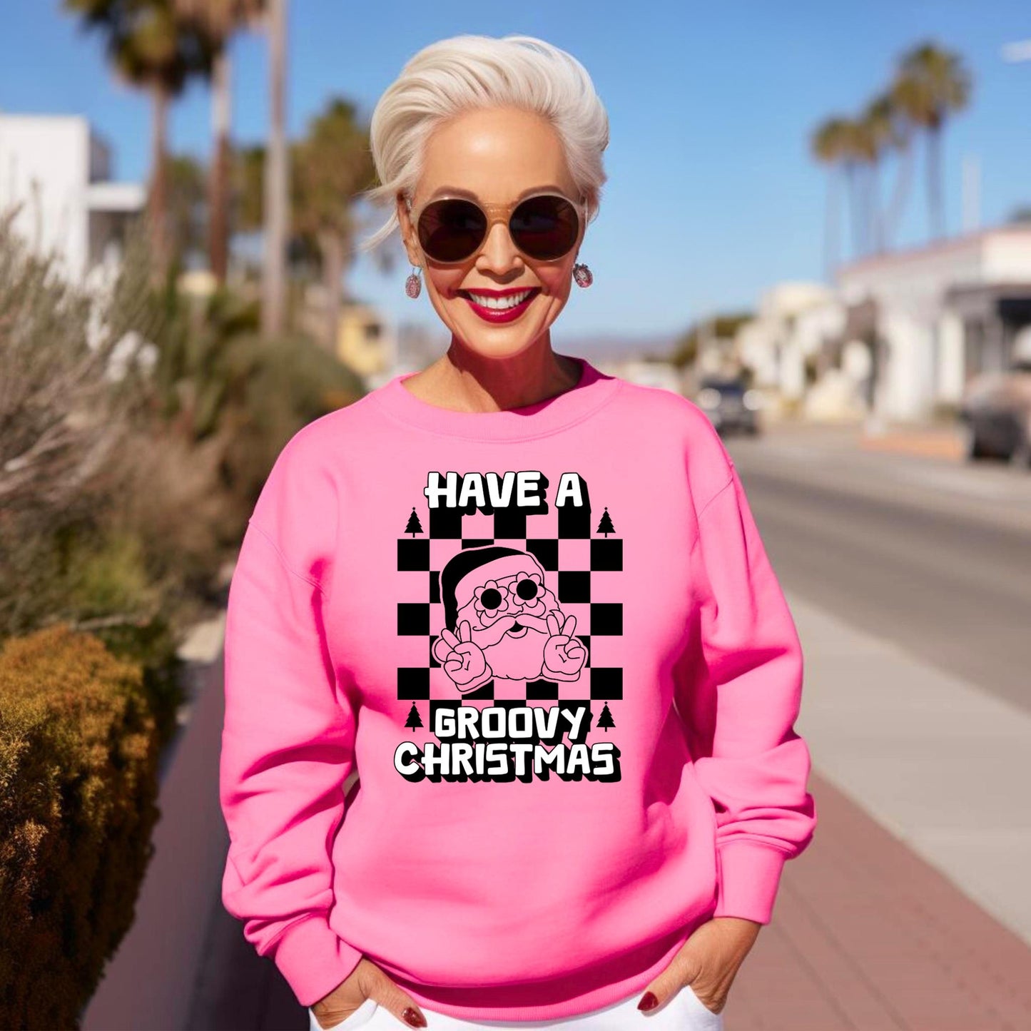 Groovy Santa Sweatshirt in Safety Pink and Safety Green, printed on a cozy Gildan 18000 crewneck. Retro Santa wears daisy sunglasses and gives a peace sign, with a bold black checkerboard background and white block text that reads “Have a Groovy Christmas.” Perfect for retro Christmas outfits, holiday party looks, matching family sweatshirts, or a unique Christmas gift idea. Unisex sizes S–5XL make it versatile for everyone.