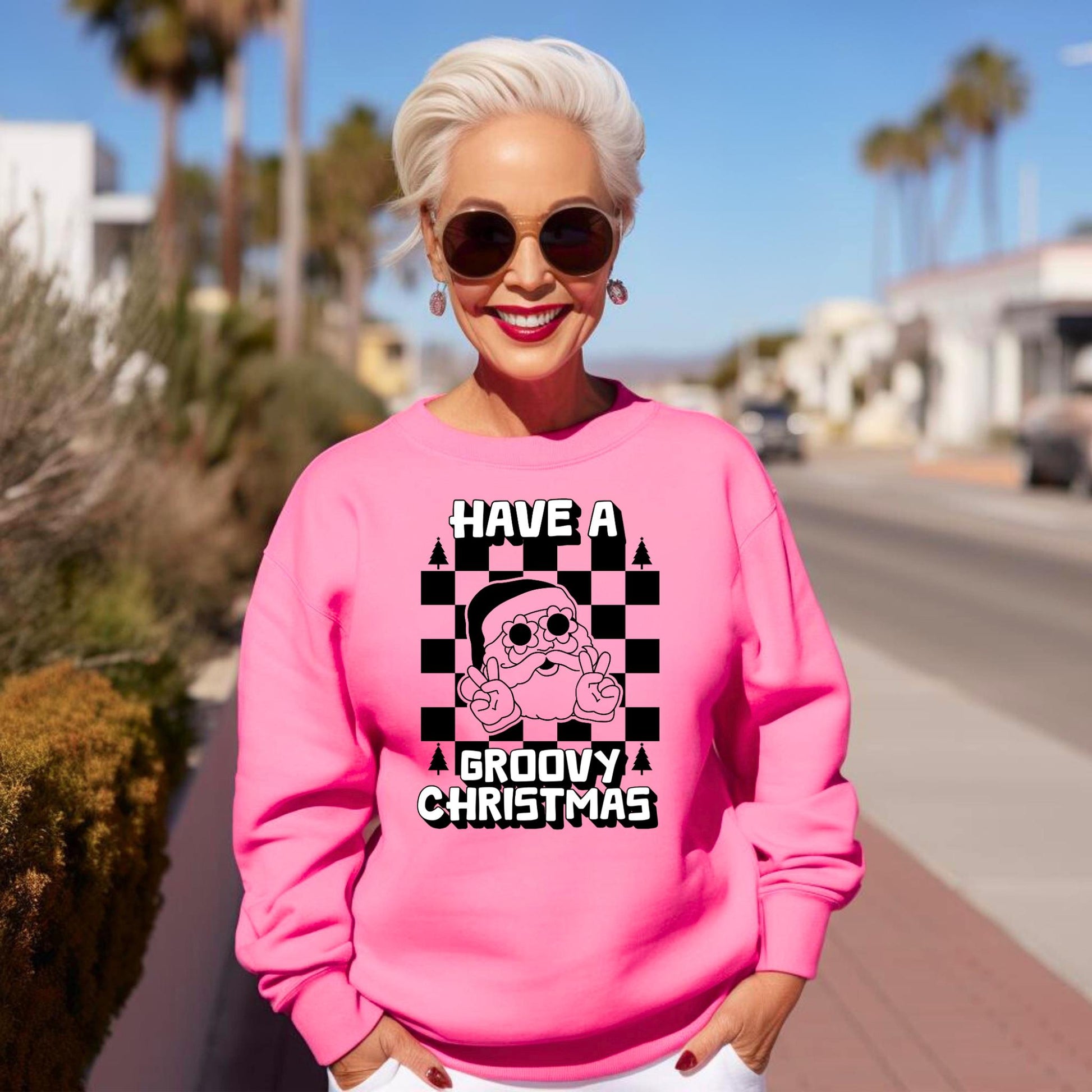 Groovy Santa Sweatshirt in Safety Pink and Safety Green, printed on a cozy Gildan 18000 crewneck. Retro Santa wears daisy sunglasses and gives a peace sign, with a bold black checkerboard background and white block text that reads “Have a Groovy Christmas.” Perfect for retro Christmas outfits, holiday party looks, matching family sweatshirts, or a unique Christmas gift idea. Unisex sizes S–5XL make it versatile for everyone.