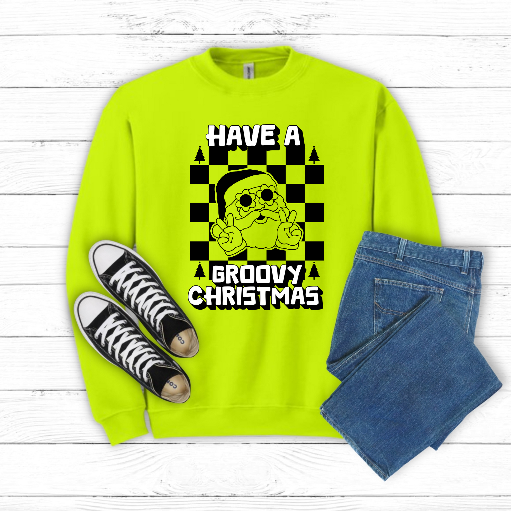 Vintage-inspired Groovy Santa Sweatshirt with retro Santa wearing flower sunglasses and throwing up a peace sign, surrounded by a checkerboard background. Bold block text reads “Have a Groovy Christmas,” making this colorful holiday pullover stand out. Available in Safety Pink and Safety Green, sizes S–5XL. Cozy crewneck makes the perfect Christmas gift, fun holiday outfit, or retro-inspired winter staple for festive family photos.