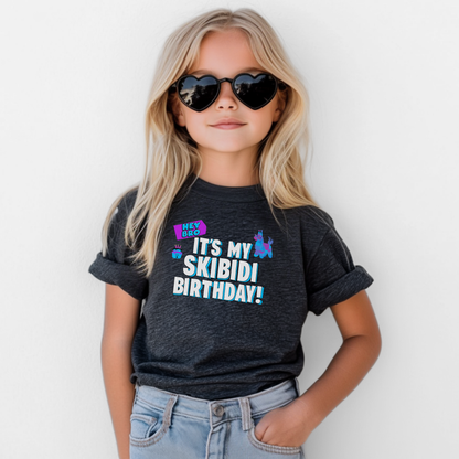 A young girl with blonde hair and heart-shaped sunglasses wears a dark charcoal heather graphic t-shirt that reads 'IT'S MY SKIBIDI BIRTHDAY!' in white and light blue, paired with light blue jeans against a plain white background. Funny kids birthday shirt featuring a Skibidi birthday design with Hey Bro It’s My Skibidi Birthday lettering, colorful graphics, and bold blue details on a comfortable black youth graphic tee